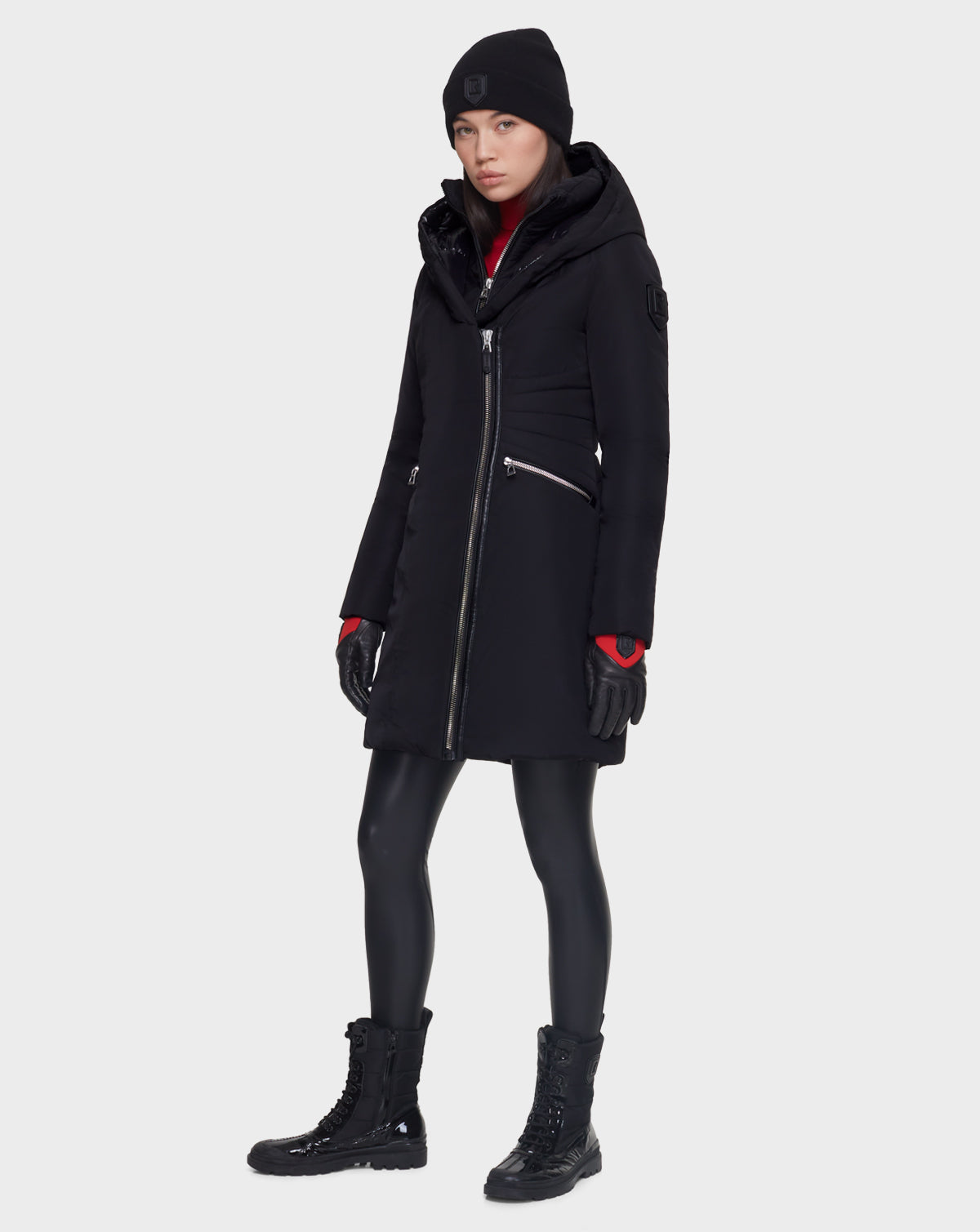 A woman wears the Rudsak CORA - L121526 BLACK women’s fitted parka made from recycled fabric, paired with a black beanie, gloves, shiny leggings, and lace-up boots, standing against a plain light gray background.
