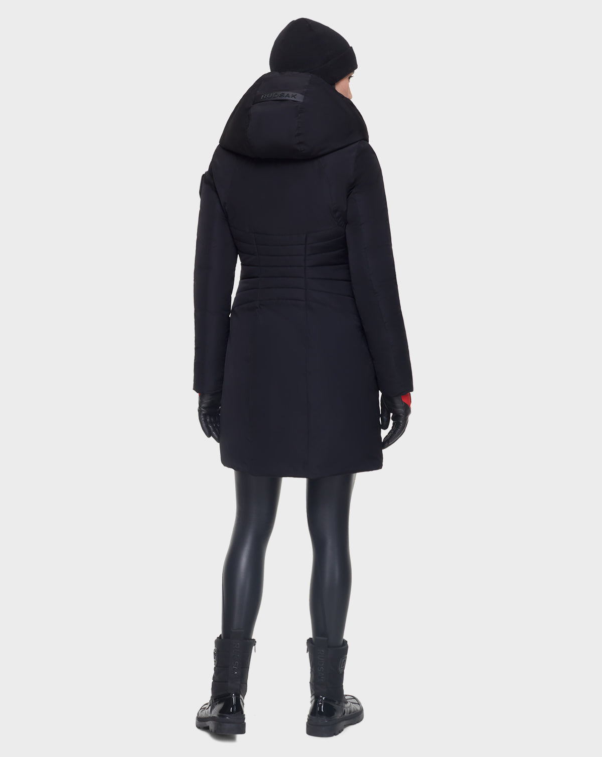 A person stands facing away, wearing Rudsak’s CORA - L121526 BLACK women’s fitted parka made with RDS-certified down, along with black leggings, boots, gloves, and a beanie against a plain white background.