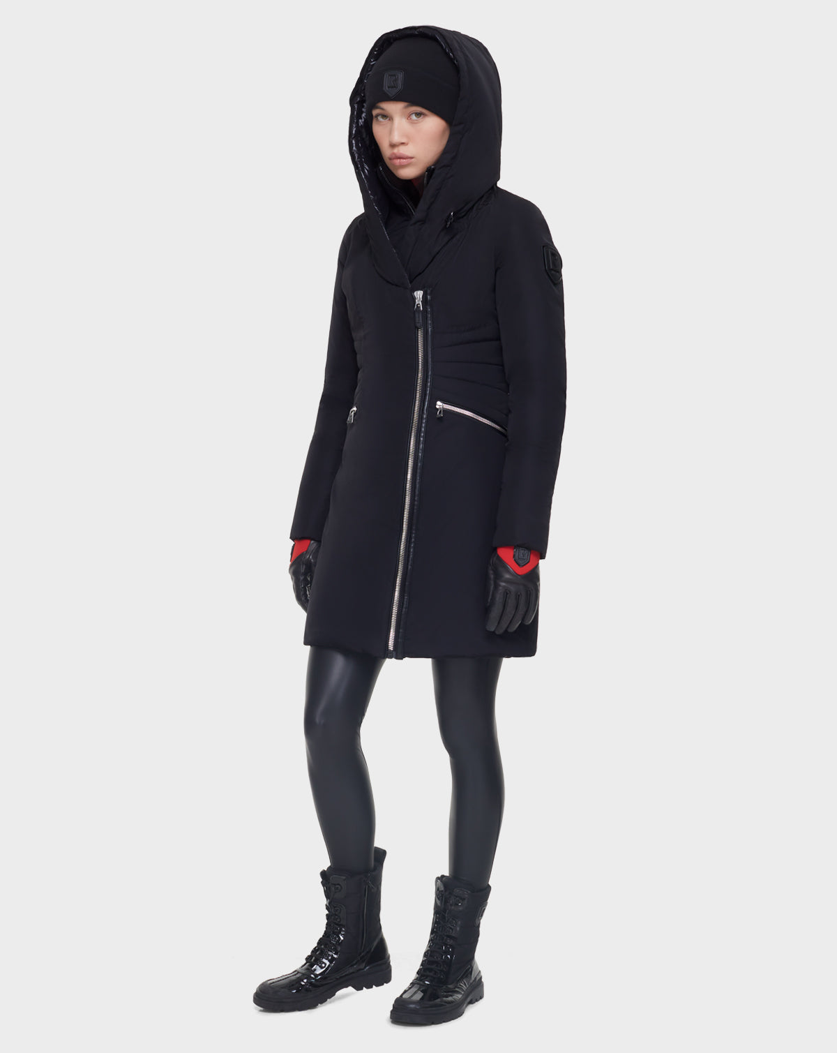 A woman wearing the Rudsak CORA - L121526 BLACK down fitted parka, black beanie, gloves, leggings, and lace-up boots stands facing forward against a plain light background.