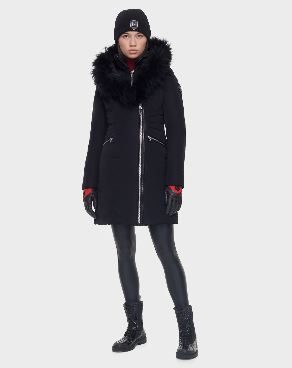 A woman faces forward in a Rudsak CORA - L121526 BLACK women’s fitted parka with a large faux fur collar, black beanie, gloves, leggings, and lace-up boots against a plain white background.