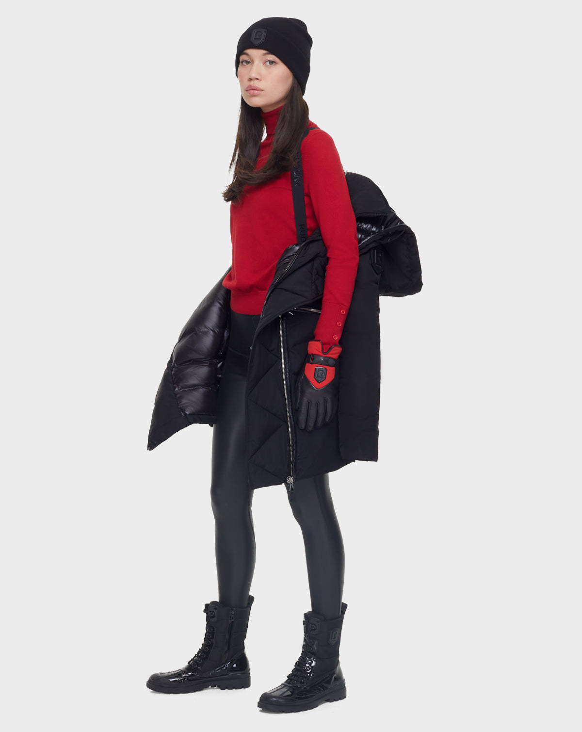 A woman in a black beanie, red sweater, gloves, shiny black leggings, and boots stands slightly to the side against a plain background, holding the Rudsak CORA - L121526 BLACK women’s fitted parka with RDS-certified down over her shoulder.