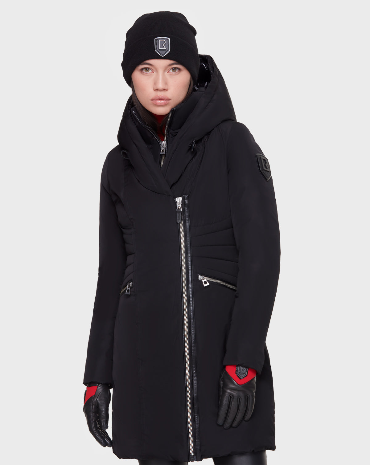 A person wears the Rudsak CORA - L121526 BLACK womens fitted parka with an asymmetrical zipper, black beanie, and gloves. Red cuff accents add a bold touch against a plain white background.