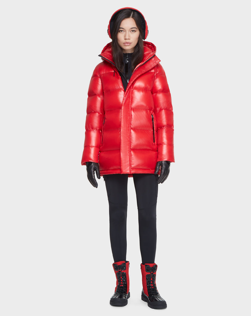 A woman faces forward in a shiny red Rudsak JOJO - 8121521 POPPY RED women’s down puffer jacket with RDS-certified down and a hood, black gloves, black pants, and red-and-black lace-up boots against a plain white background.