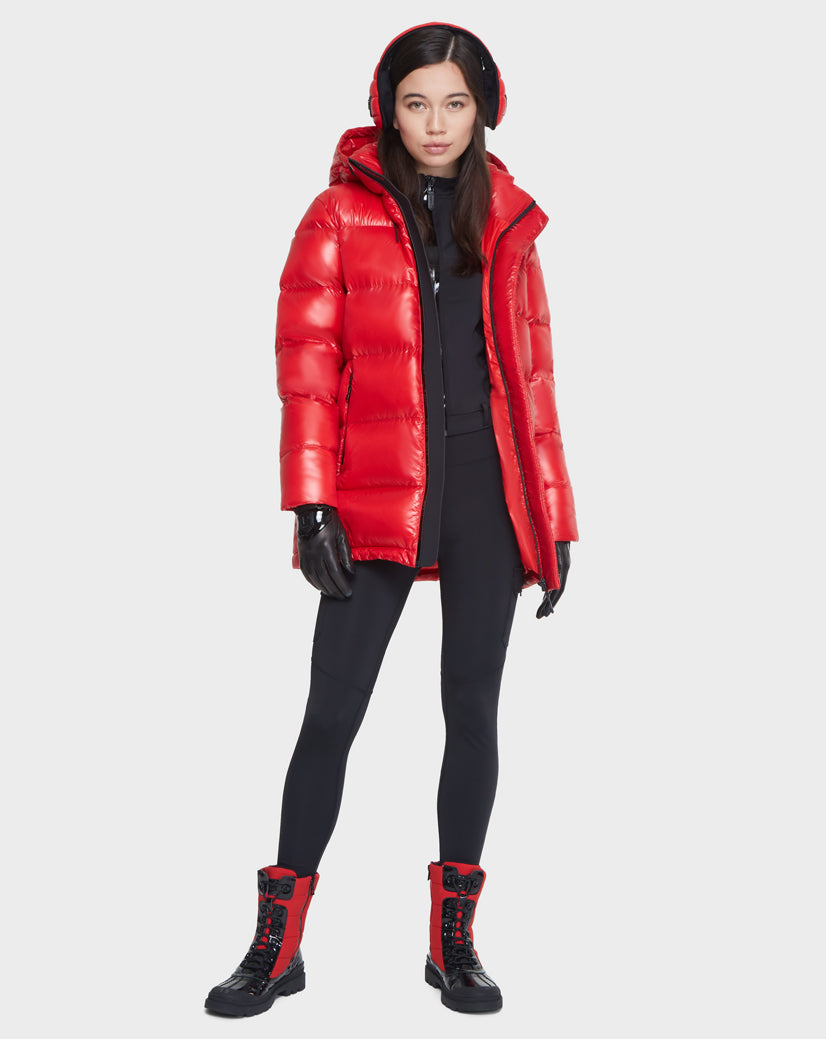 A woman wears the Rudsak JOJO - 8121521 POPPY RED down puffer jacket with black gloves, earmuffs, fitted black pants, and black-and-red lace-up boots, standing facing forward against a plain light background.