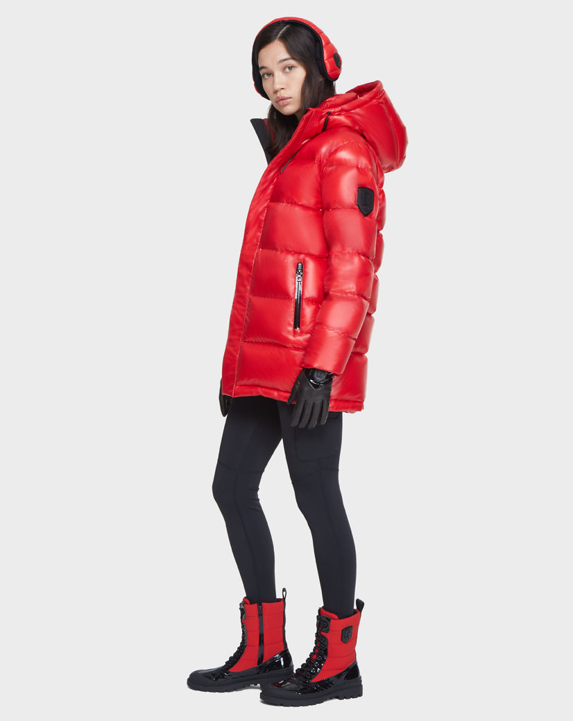 A woman stands sideways against a white background wearing the Rudsak JOJO - 8121521 POPPY RED down puffer jacket with a hood, paired with red earmuffs, black gloves, leggings, and black boots with red accents.