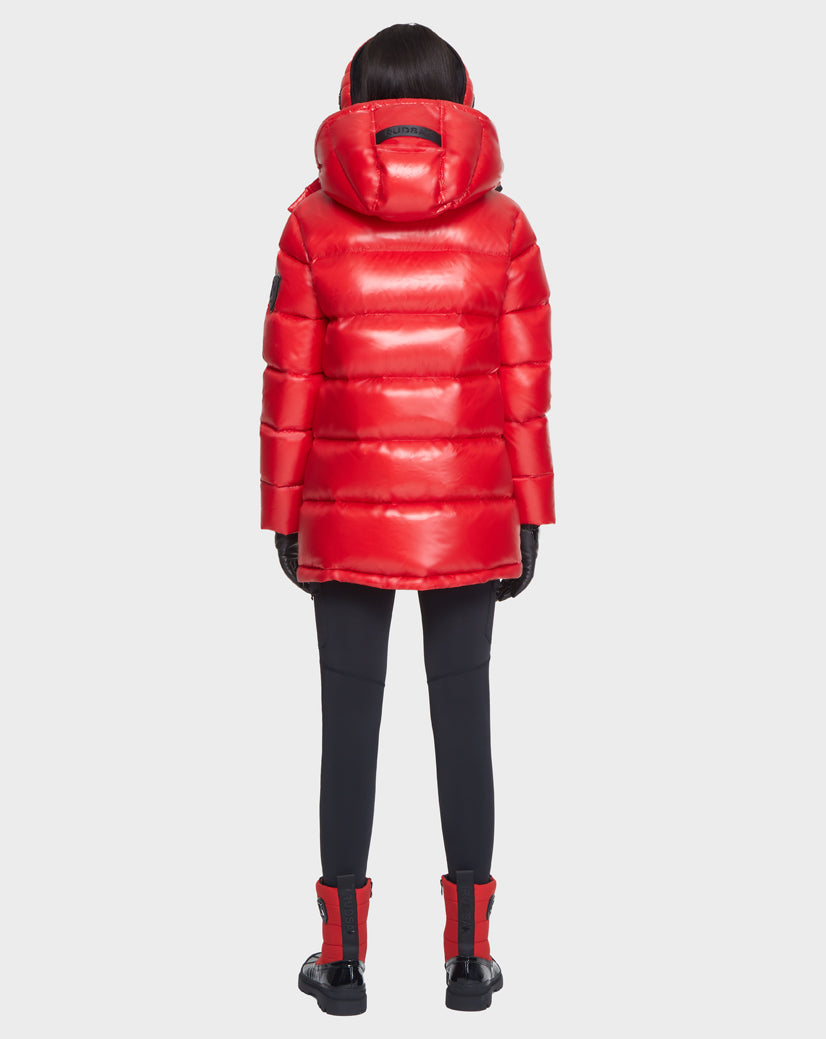 A person stands facing away, wearing the Rudsak JOJO - 8121521 POPPY RED women's down puffer jacket with a hood, paired with black gloves, fitted black pants, and red and black boots against a plain light background.