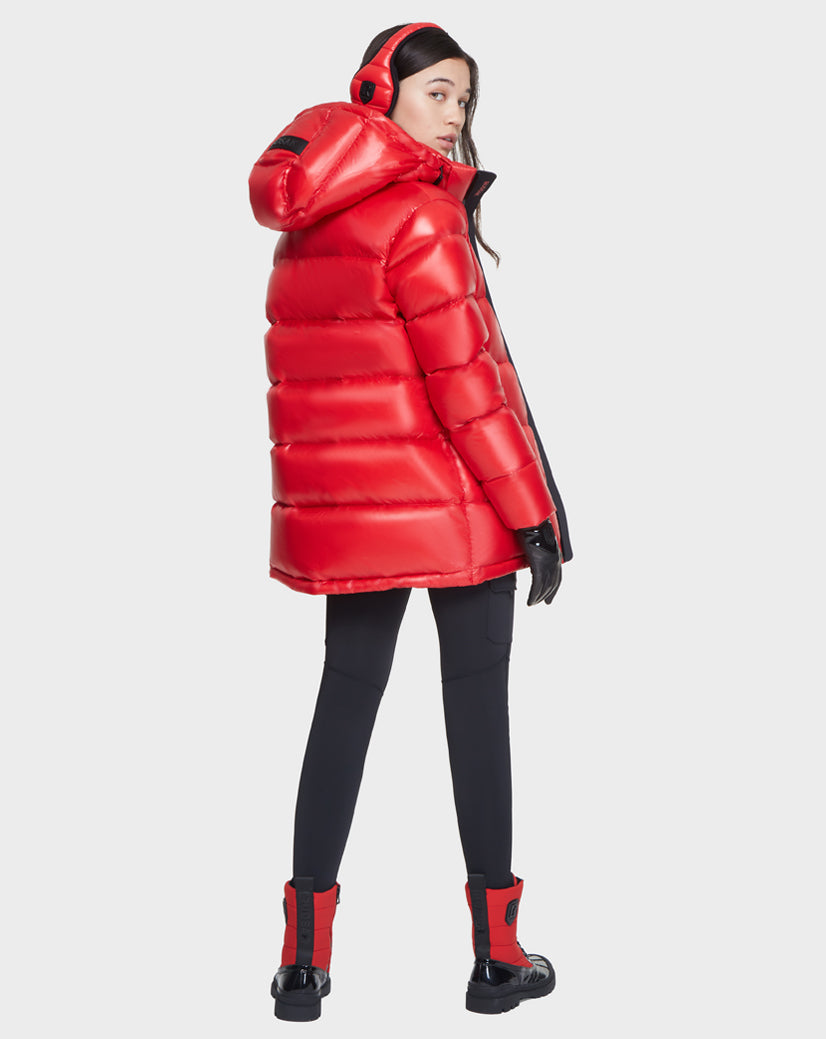 A woman wearing the Rudsak JOJO - 8121521 POPPY RED down puffer jacket with a hood, paired with black leggings, gloves, red earmuffs, and red-and-black boots stands facing away and looks over her shoulder.