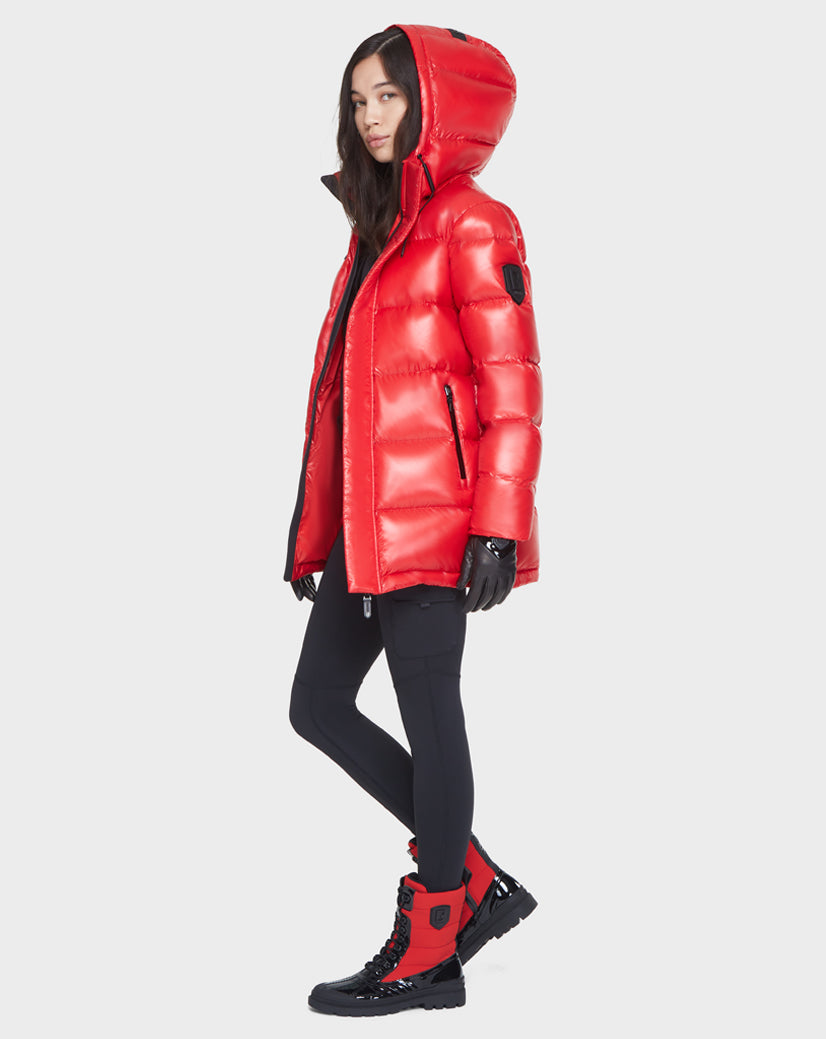 A woman stands sideways against a plain light background, wearing the Rudsak JOJO - 8121521 POPPY RED women's down puffer jacket, paired with black gloves, black leggings, and red and black boots.