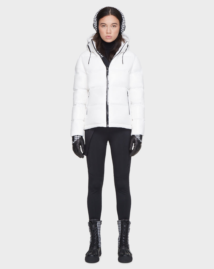 A woman faces forward wearing the Rudsak JUNO - 8121553 OPTIC WHITE water-repellent, RDS-certified down puffer jacket with hood, black leggings, gloves, and boots, creating a chic monochrome winter look against a plain light background.