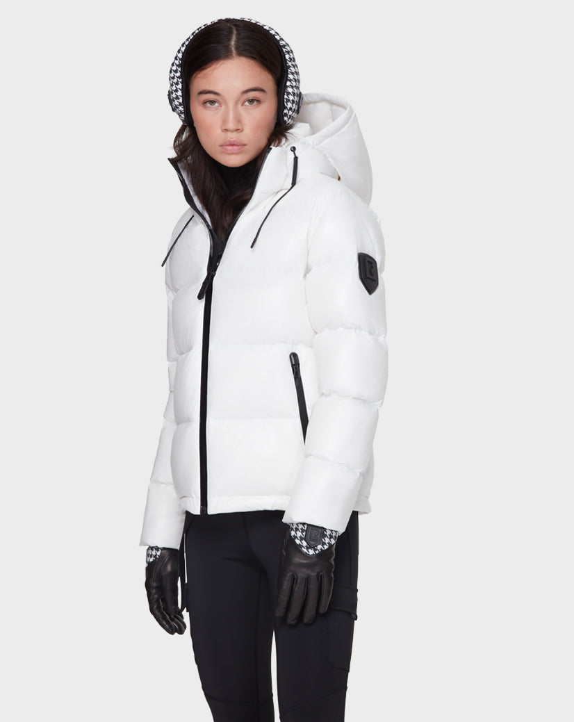 A woman in the Rudsak JUNO - 8121553 OPTIC WHITE water-repellent puffer jacket with a hood stands against a light grey background, paired with black gloves, black pants, and checkered earmuffs, facing slightly to the side.