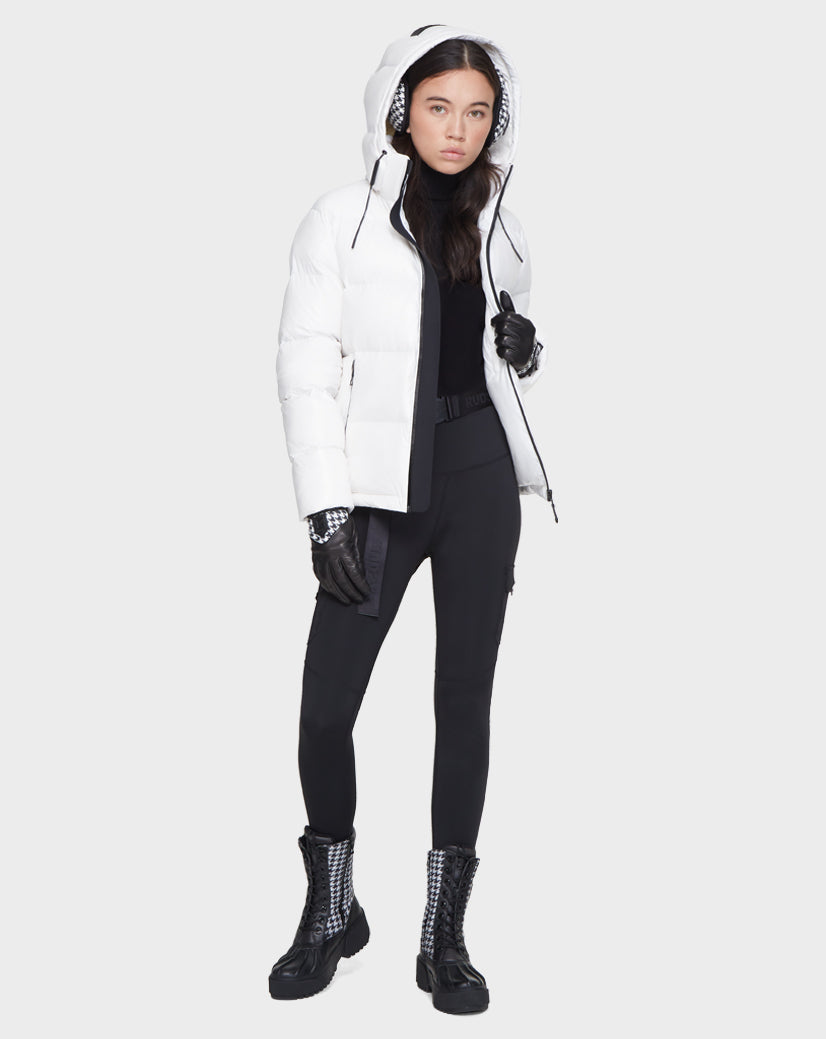 A woman stands against a light background wearing the Rudsak JUNO - 8121553 OPTIC WHITE, a water-repellent women’s puffer jacket with the hood up, paired with black gloves, leggings, top, and checkered black boots.