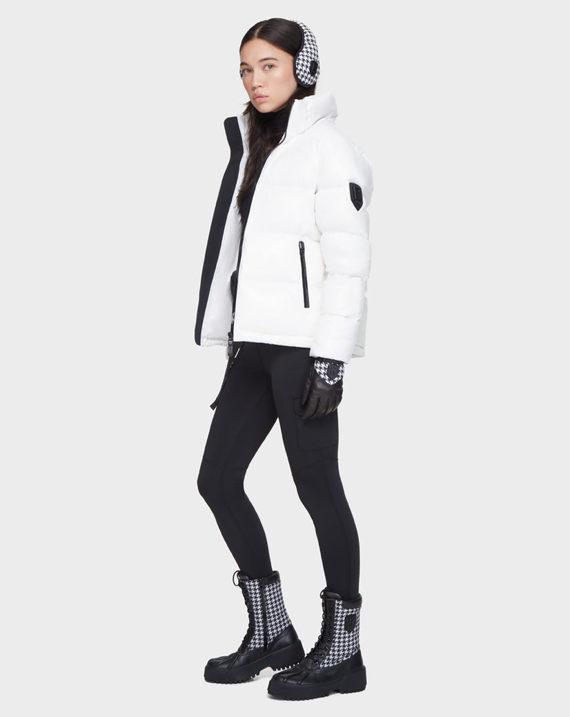 A woman wears the Rudsak JUNO - 8121553 OPTIC WHITE puffer jacket, black leggings, houndstooth boots, checkered gloves, and earmuffs as she stands against a plain light gray background.