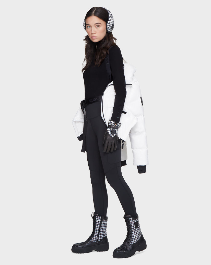 A woman stands against a light grey background wearing Rudsak's JUNO - 8121553 OPTIC WHITE women’s puffer jacket draped over her shoulders, along with black leggings, a black top, gloves, checkered earmuffs, and boots.