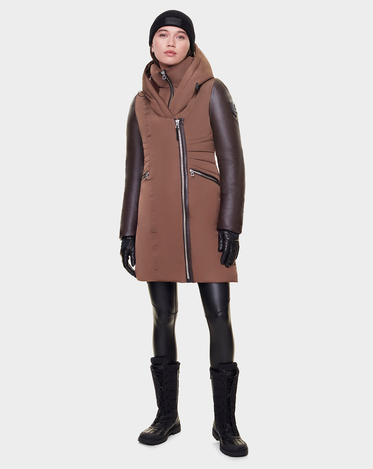 A woman wearing the Rudsak MISA - L121506 CAOCAO FAUX FUR brown sustainable winter coat with asymmetrical zipper, black leather sleeves, black gloves, leggings, boots, and beanie stands facing forward against a plain light background.