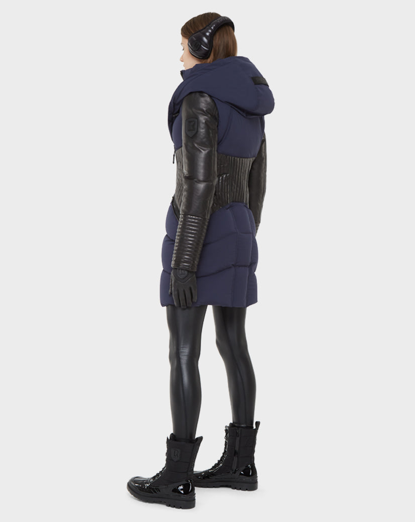 A person stands at an angle against a plain background, wearing the Rudsak SHAUNA - 8121509 NAVY BLUE RDS-certified down padded vest with hood, black leather sleeves, gloves, leggings, lace-up boots, and earmuffs.