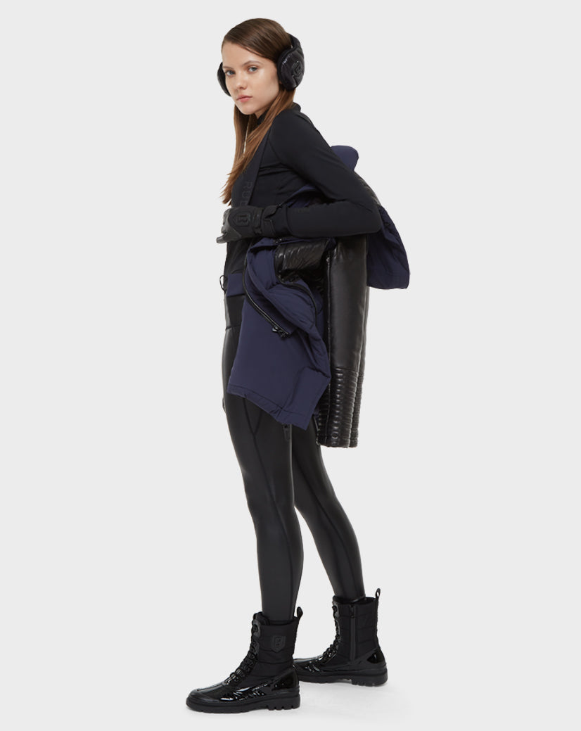 A woman in winter gear—black earmuffs, gloves, long-sleeve top, shiny leggings, and lace-up boots—holds the Rudsak SHAUNA - 8121509 NAVY BLUE quilted down puffer and a black jacket against a plain light background.