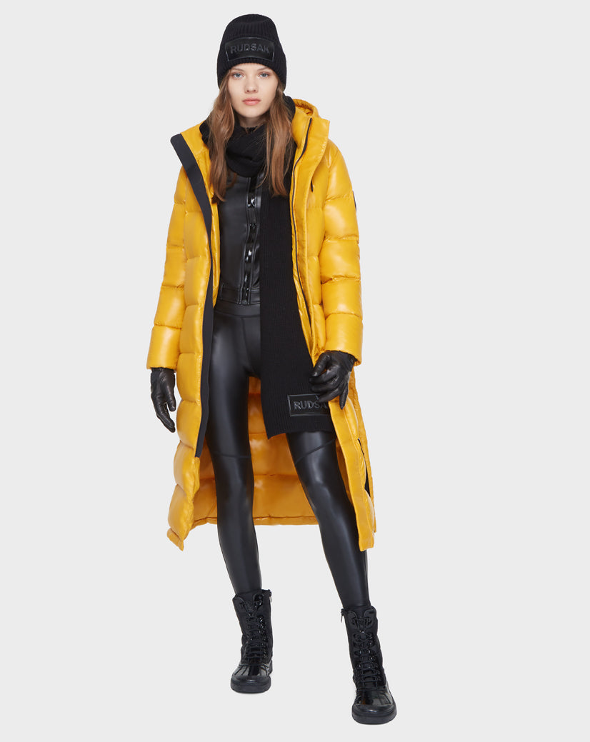 A woman stands against a light gray background wearing the Rudsak MAO - 8121506 YELLOW shiny down long puffer coat over black faux leather leggings, a black jacket, combat boots, gloves, scarf, and a beanie.