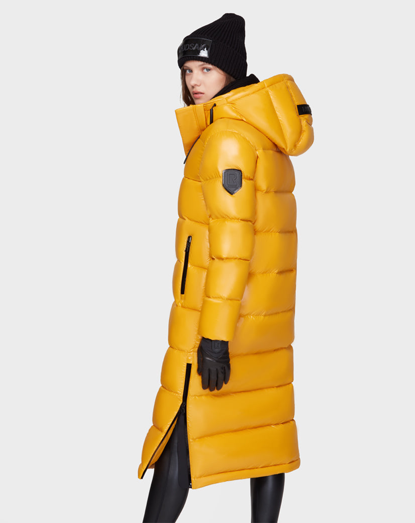 A person in the Rudsak MAO - 8121506 YELLOW long puffer coat with RDS-certified down, styled with a black beanie, gloves, and leggings, stands sideways against a plain light background.