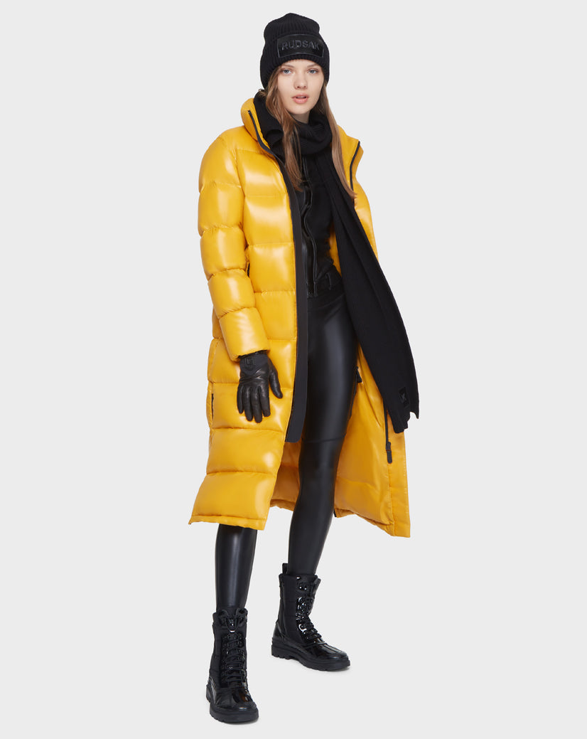 A woman wearing the Rudsak MAO - 8121506 YELLOW long puffer coat with a black beanie, scarf, gloves, shiny black lace-up boots, and leggings stands against a plain light background.
