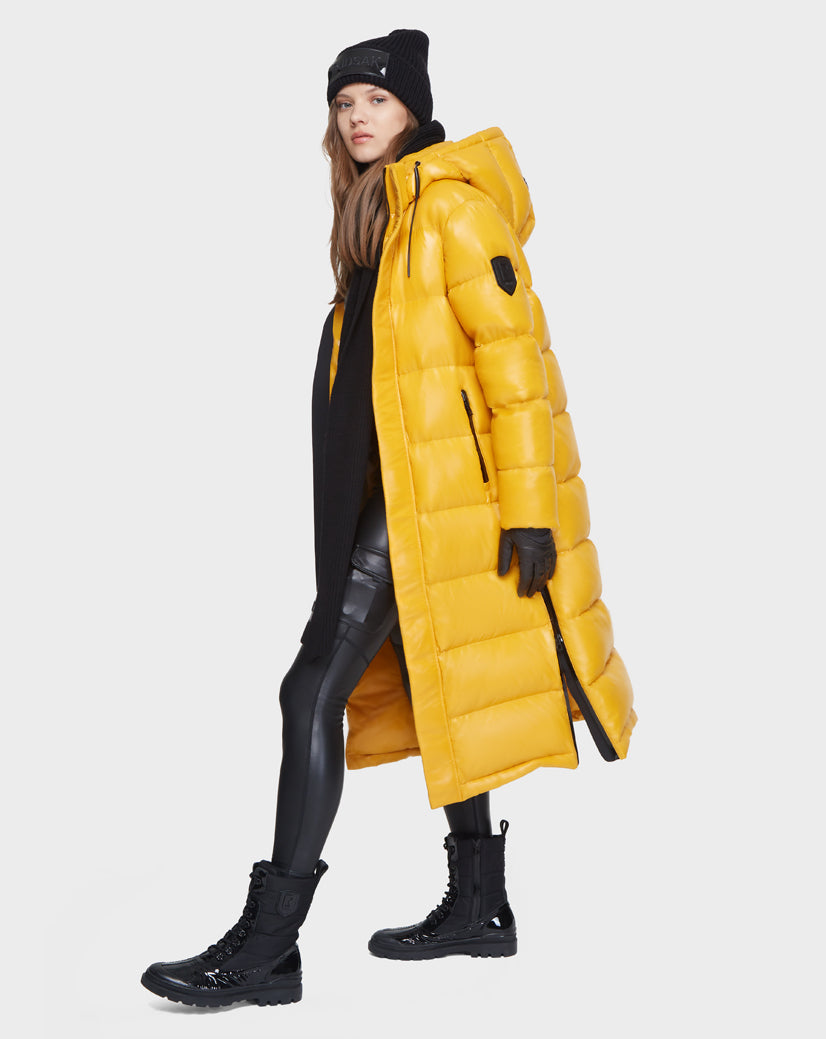 A person wearing the Rudsak MAO - 8121506 YELLOW long, RDS-certified down puffer coat, plus black winter accessories and shiny leggings with combat boots, stands against a plain white background.
