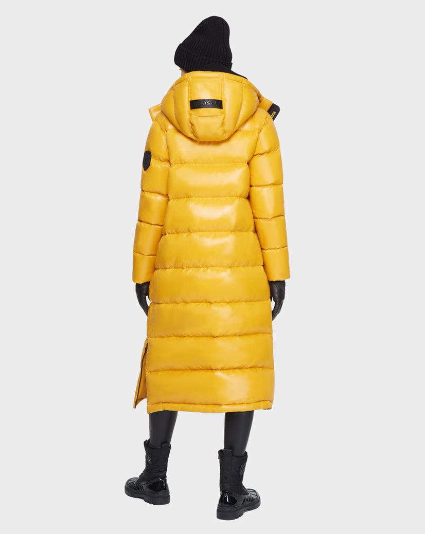 A person faces away, wearing the Rudsak MAO - 8121506 YELLOW shiny long hooded puffer coat with black gloves, beanie, pants, and boots against a plain light background.