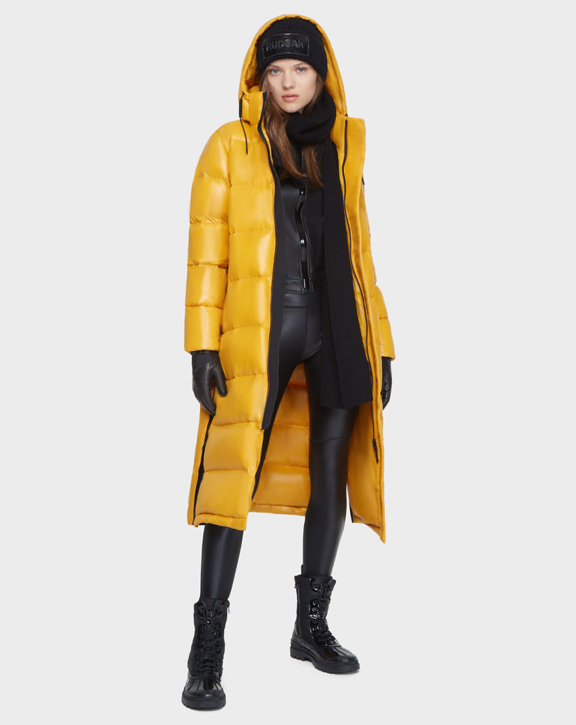 A woman wearing the Rudsak MAO - 8121506 YELLOW long puffer coat, filled with RDS-certified down, pairs it with a black beanie, scarf, gloves, leggings, and lace-up boots against a plain white background.