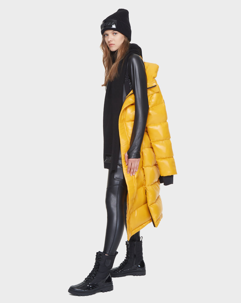 A woman in a black outfit and boots stands sideways against a white background, holding the Rudsak MAO - 8121506 YELLOW women's down puffer draped over one shoulder.