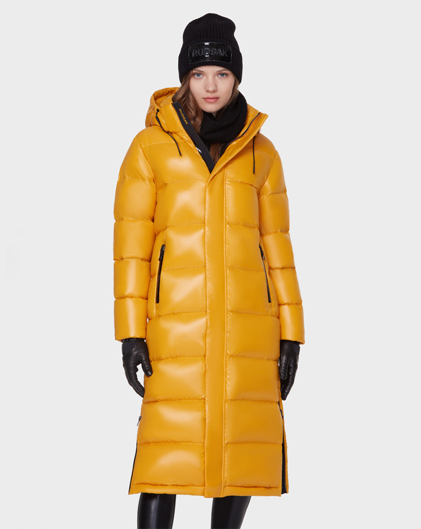 A woman wears the MAO - 8121506 YELLOW long puffer coat by Rudsak, filled with RDS-certified down, paired with a black beanie, gloves, and pants as she stands against a plain white background.