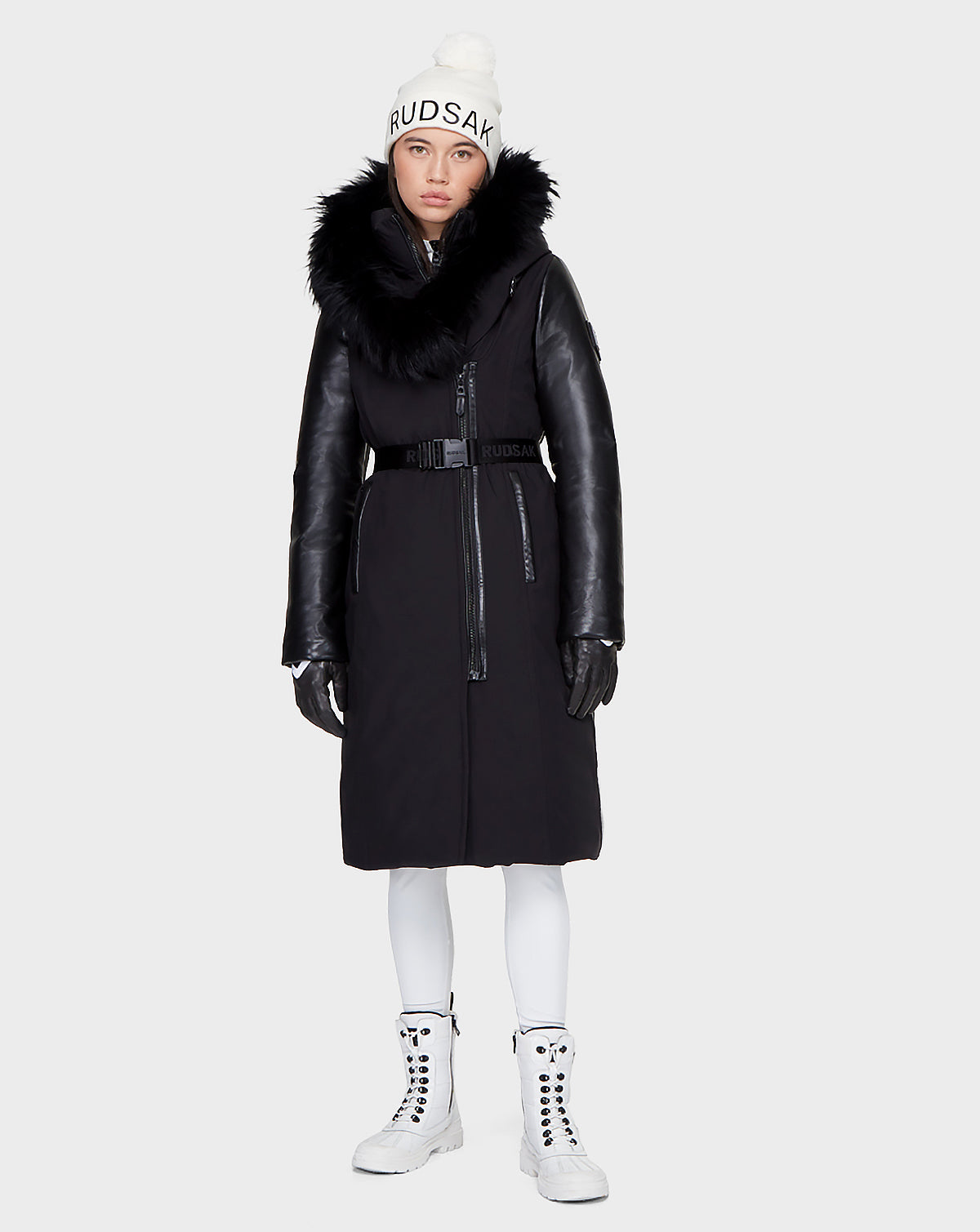 A woman faces forward wearing the Rudsak JANIS - 8121532 black long down parka with RDS-certified down, a fur-trimmed hood, black gloves, white leggings and boots, and a white beanie with "RUDSAK" on it.