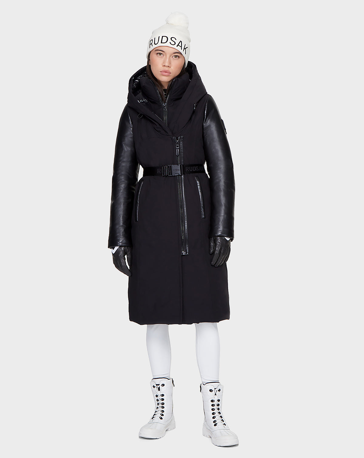 A woman faces forward wearing the Rudsak JANIS - 8121532 BLACK women’s winter coat with leather sleeves, black gloves, a white Rudsak beanie, white leggings, and white lace-up boots against a plain light background.
