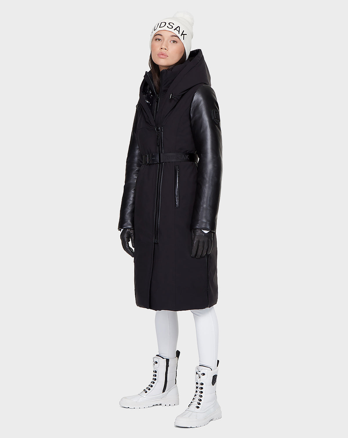 A woman stands against a plain light background wearing the Rudsak JANIS - 8121532 BLACK down parka with leather sleeves, styled with a white knit beanie, white leggings, black gloves, and white lace-up boots.