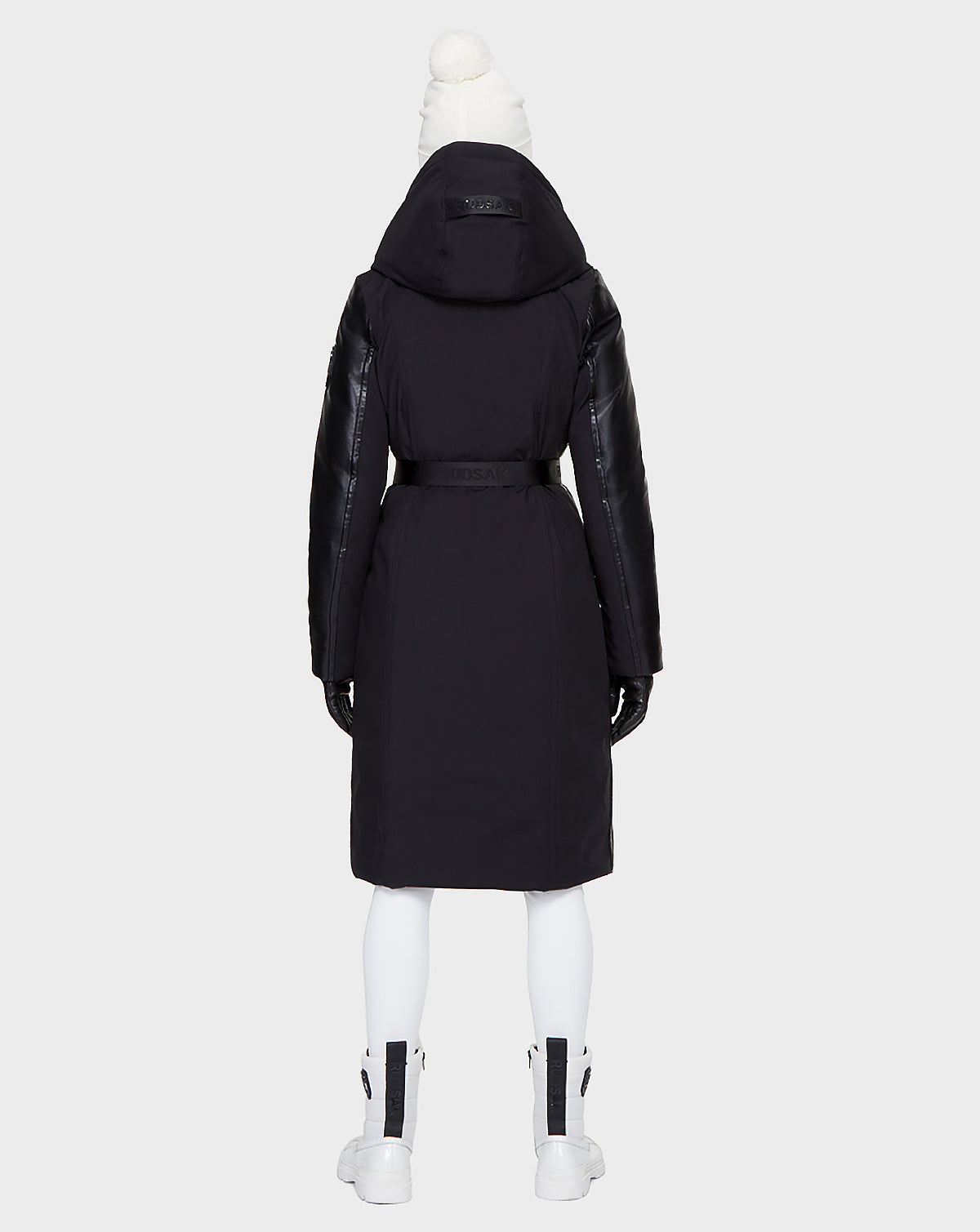 A person faces away wearing the Rudsak JANIS - 8121532 BLACK winter coat with a hood and black leather sleeves, styled with a white pom-pom hat, white leggings, black gloves, and zipped white boots against a light gray background.