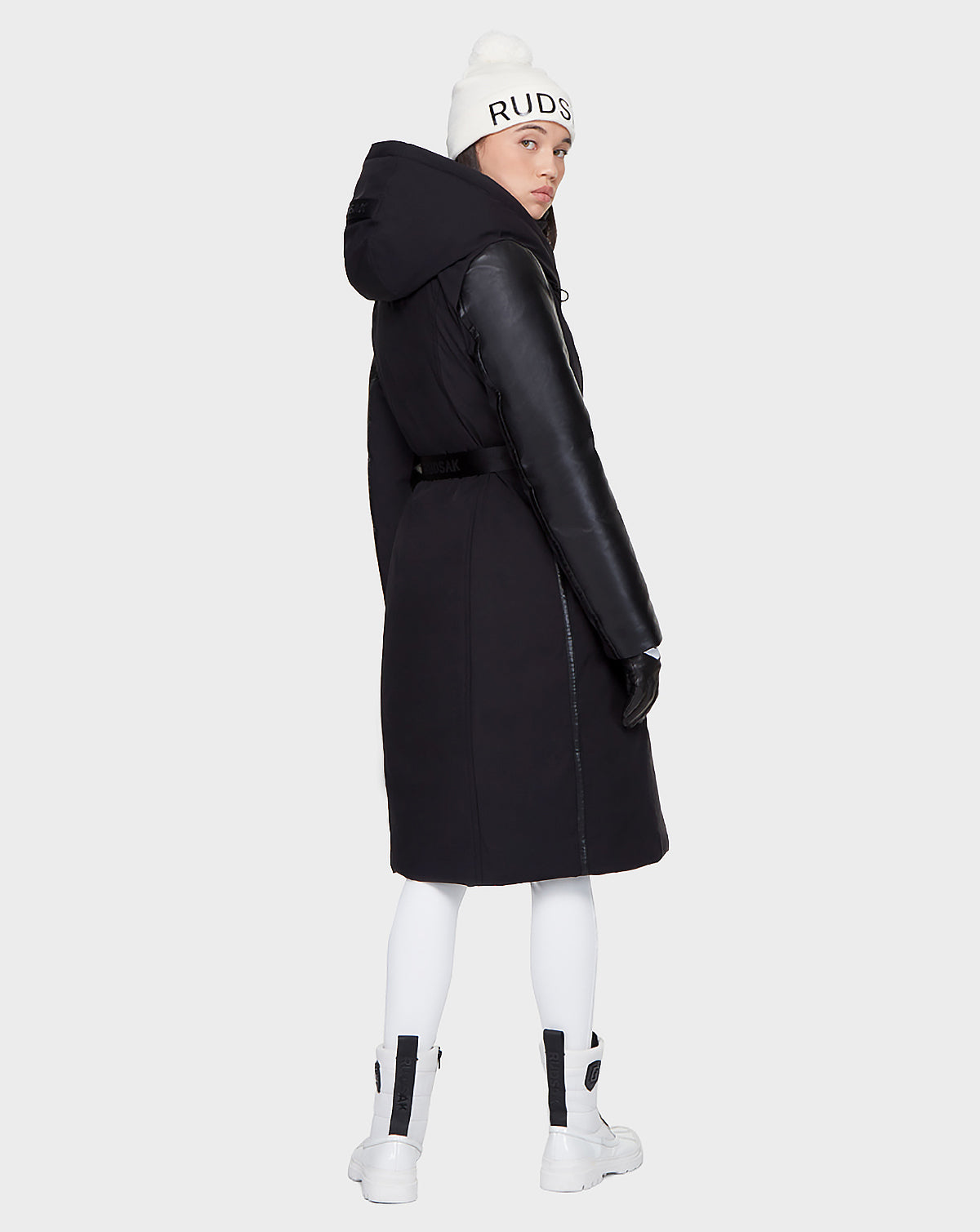 A person wears the Rudsak JANIS - 8121532 BLACK women’s winter coat, a white knit hat with "RUDSAK" text, white leggings, white boots, and black gloves, standing slightly turned and looking over their shoulder.
