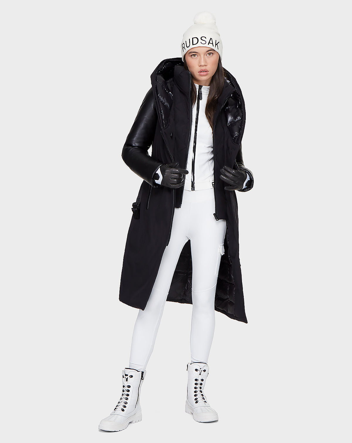 A woman in the Rudsak JANIS - 8121532 BLACK long down parka with leather sleeves, white pants, lace-up boots, a pom-pom beanie, and black gloves faces forward against a light background. The coat is filled with RDS-certified down.