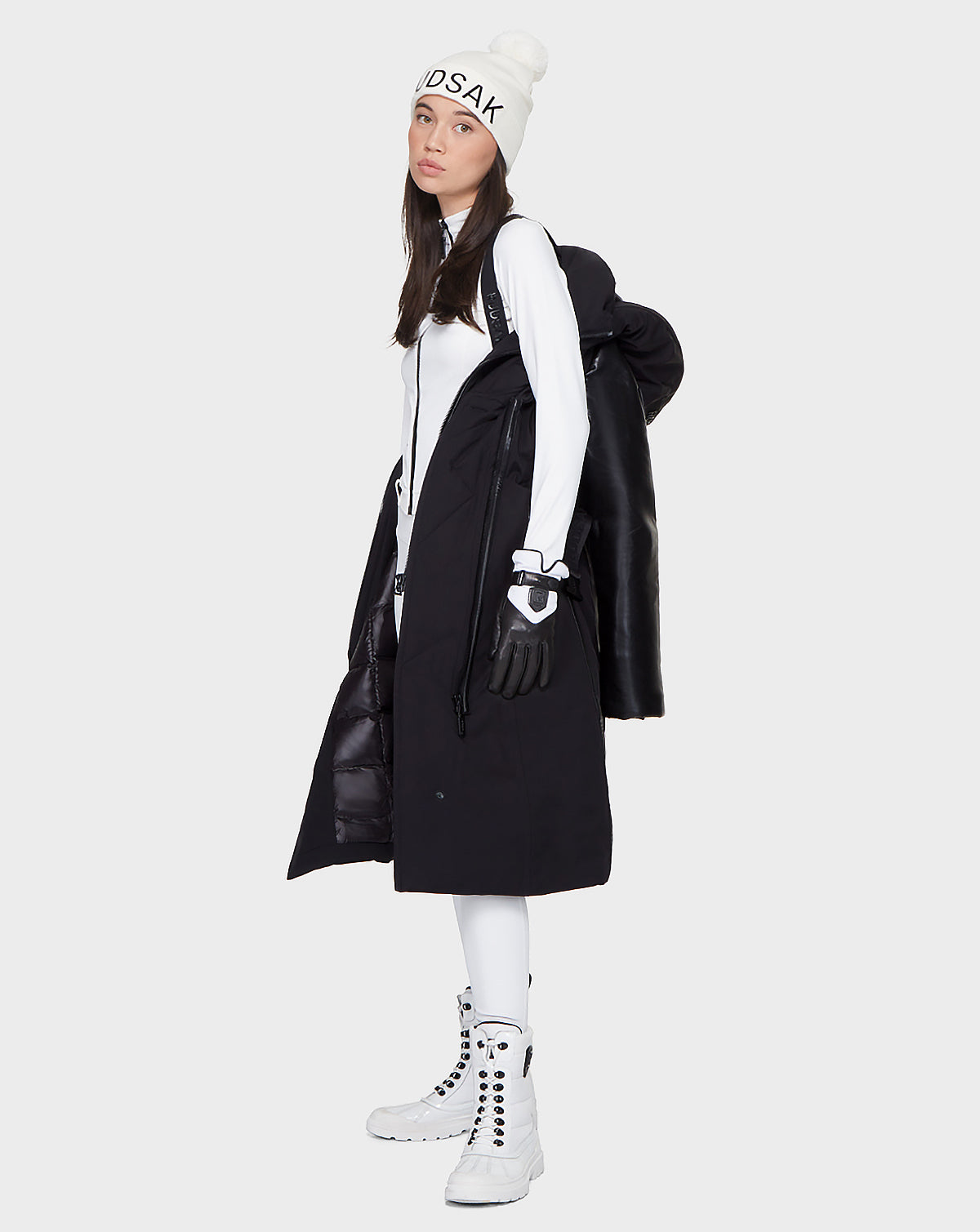 A woman wears a white beanie, turtleneck, long down parka, black gloves, white pants, and lace-up boots. She stands against a plain background with a Rudsak JANIS - 8121532 BLACK backpack and gazes slightly to the side.