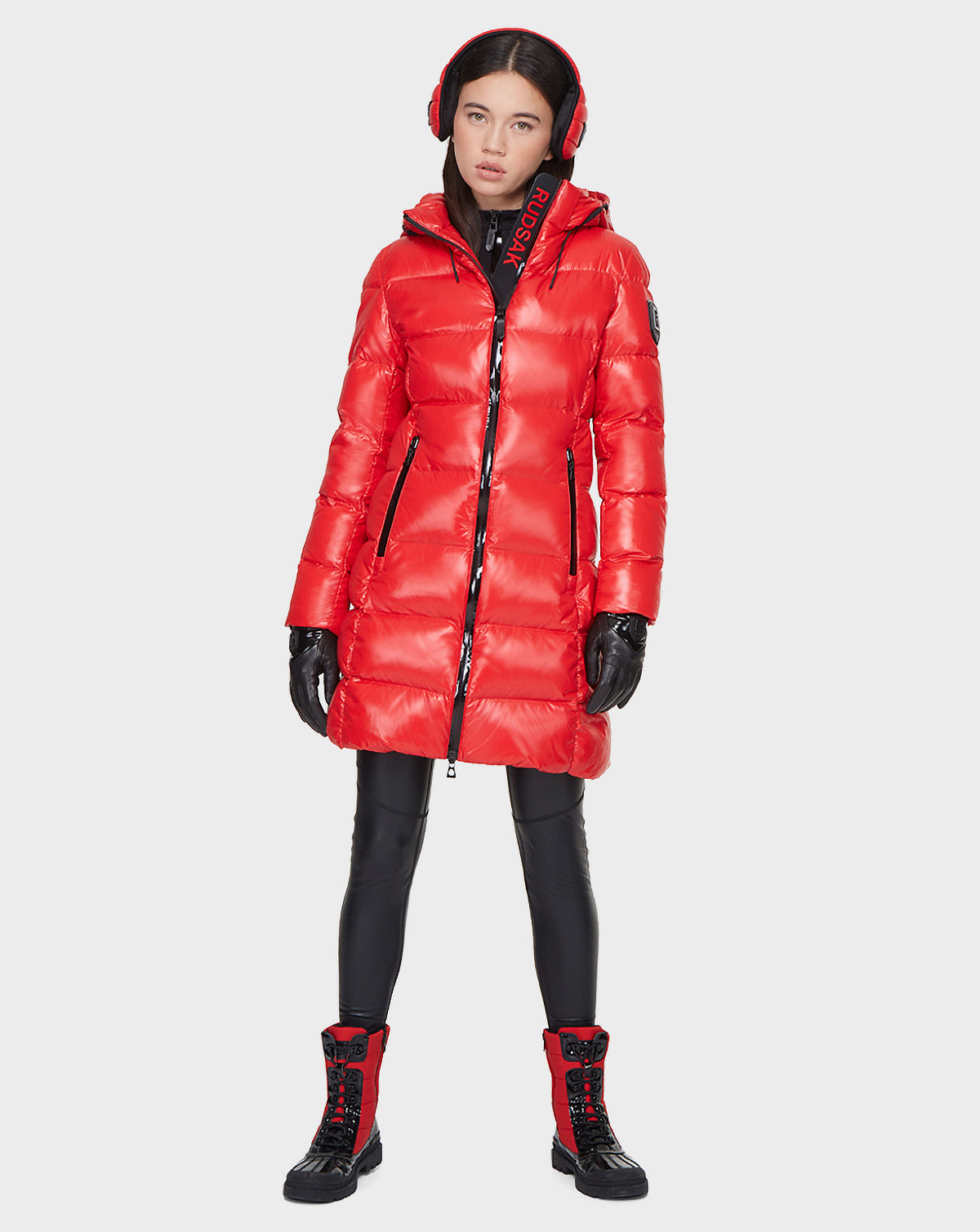 A woman stands facing forward against a plain white background, wearing the Rudsak MILIA - 8121552 POPPY RED down puffer coat with a hood, black gloves and leggings, red earmuffs, and red and black lace-up boots.