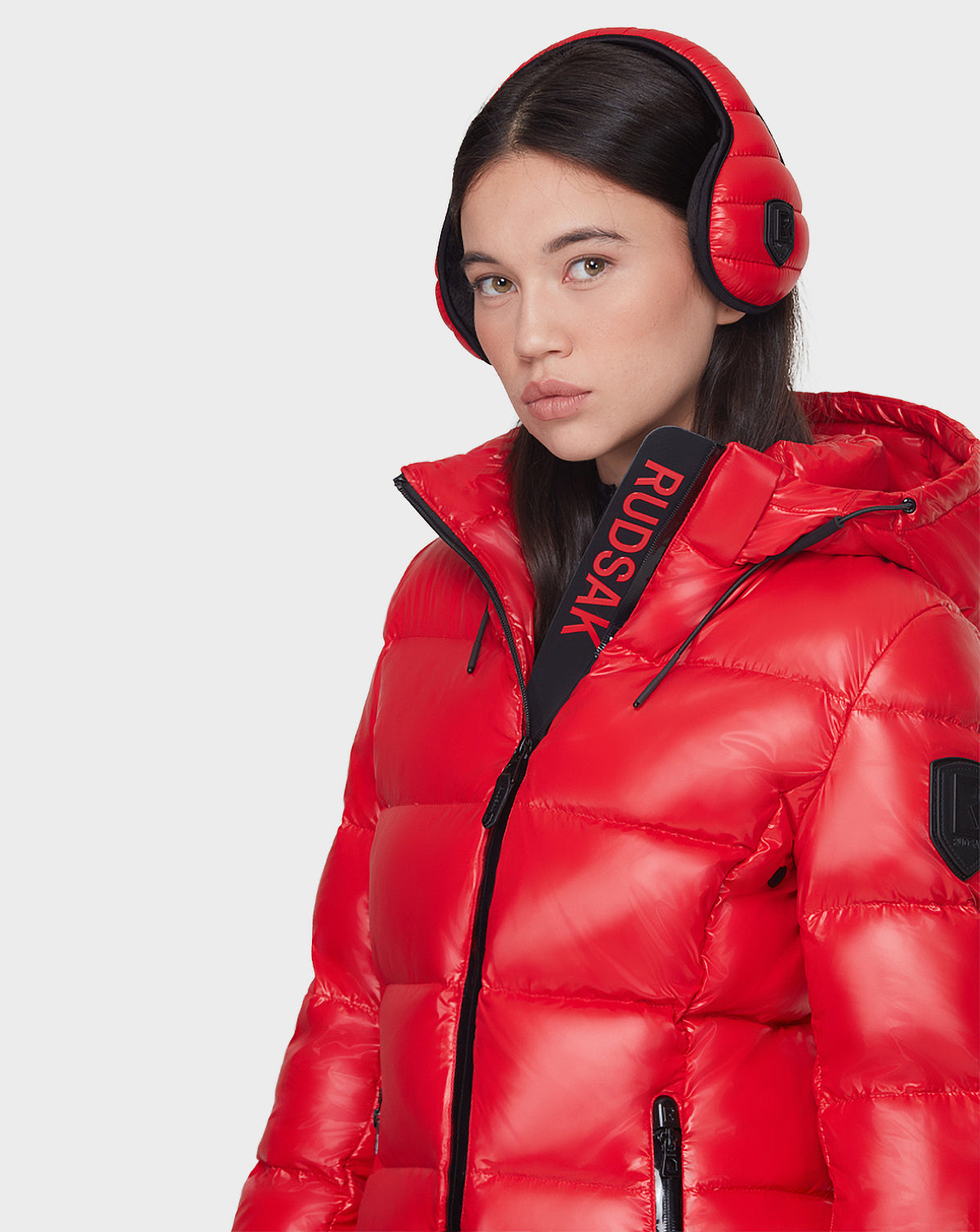 A woman with straight brown hair wears the Rudsak MILIA - 8121552 POPPY RED mid-length quilted down puffer jacket, partially zipped, with matching earmuffs and a RUDSAK-branded strap around her neck as she looks to the side.