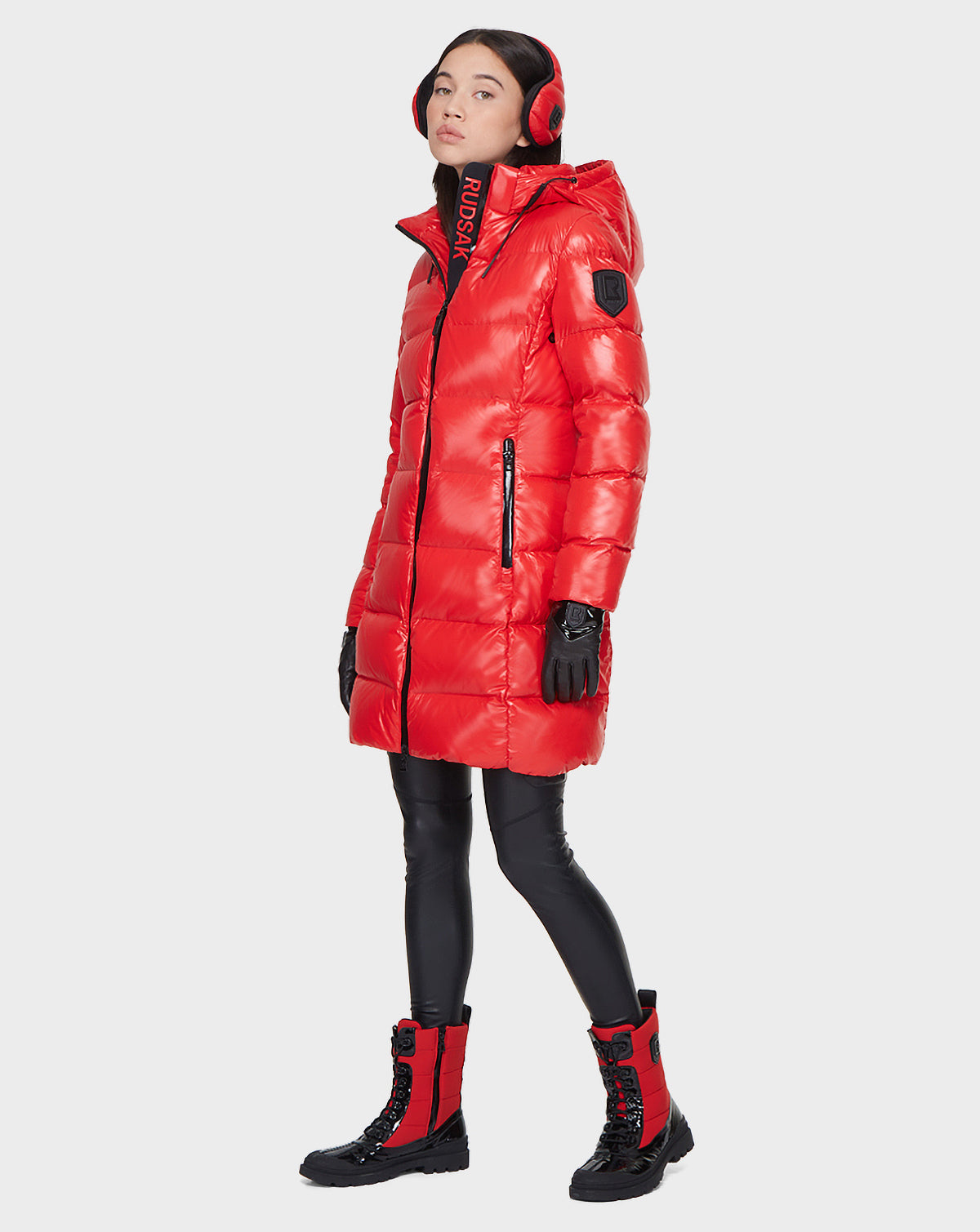 A woman wearing the Rudsak MILIA - 8121552 POPPY RED down puffer coat stands against a white background, paired with black leggings, red and black lace-up boots, black gloves, and red earmuffs.