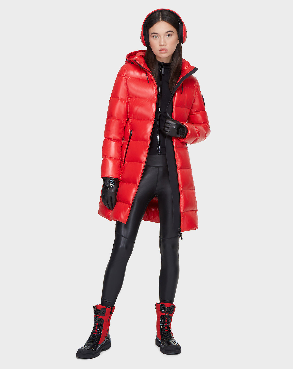 A woman faces forward, wearing the Rudsak MILIA - 8121552 POPPY RED shiny down jacket with hood, black gloves and leggings, a black zip-up top, red earmuffs, and matching red-and-black lace-up boots against a light gray background.