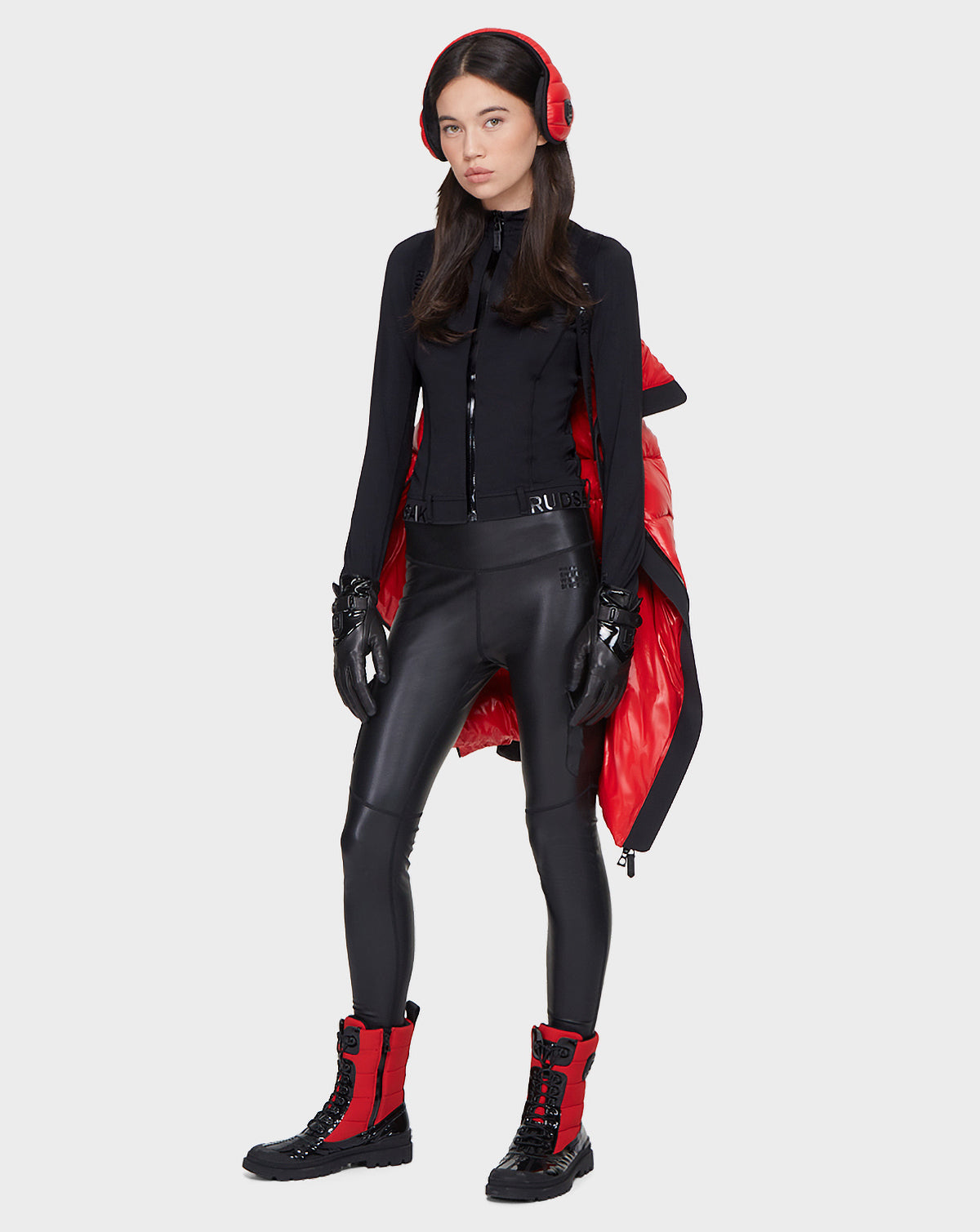 A woman faces forward in black leggings, a fitted black jacket, red-and-black boots, black gloves, and red earmuffs with a Rudsak MILIA - 8121552 POPPY RED quilted jacket draped over her shoulders. The background is plain white.