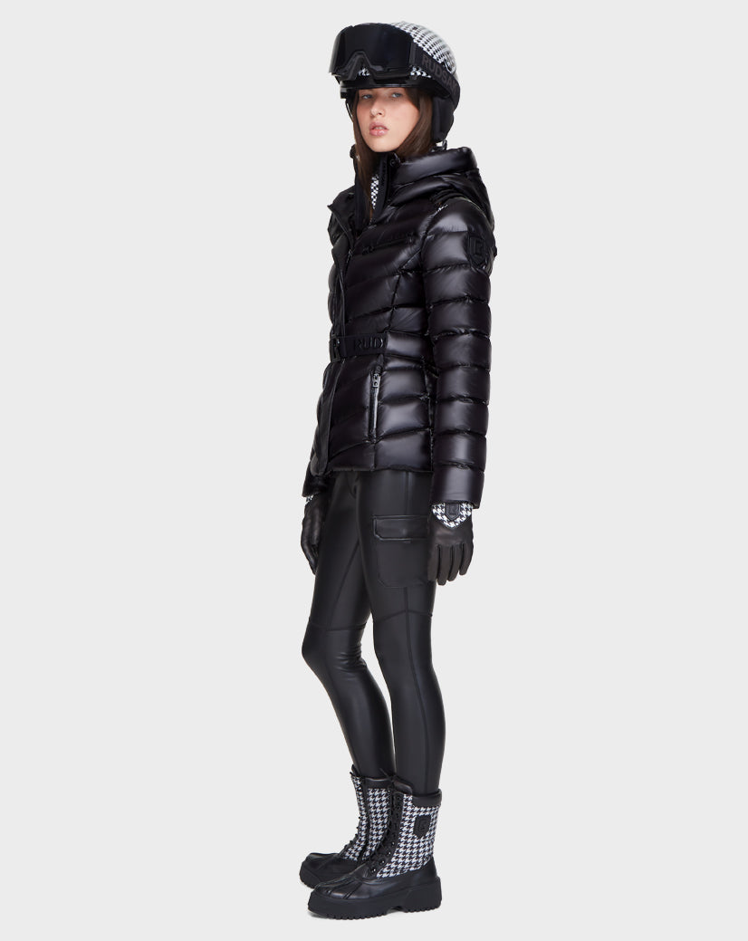 A woman stands sideways in Rudsak's MASORA - 8121535 BLACK: a sleek belted puffer ski jacket with RDS-certified down. She pairs it with fitted pants, gloves, boots, and a checkered visor helmet, gazing ahead neutrally.