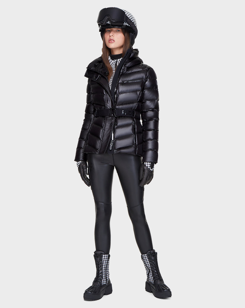 A woman wears the Rudsak MASORA - 8121535 BLACK belted puffer jacket with black faux leather leggings, lace-up boots, gloves, a patterned turtleneck, and a black visor cap with an attached checkered scarf.
