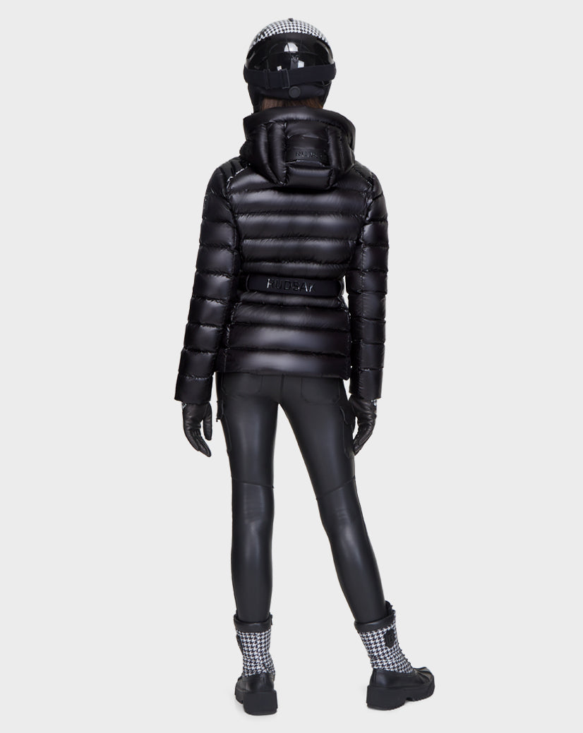 A person stands facing away, wearing the Rudsak MASORA - 8121535 BLACK women's belted puffer jacket with a hood, paired with black fitted pants, gloves, checkered cuff boots, and a black helmet with a white checkered pattern.