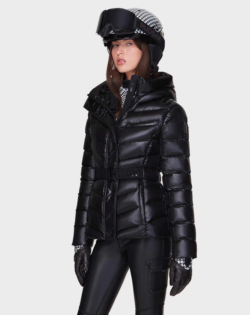 A woman wearing the Rudsak MASORA - 8121535 BLACK shiny belted puffer ski jacket, black ski pants, gloves, and a helmet with goggles stands against a plain light background, looking forward with a neutral expression.