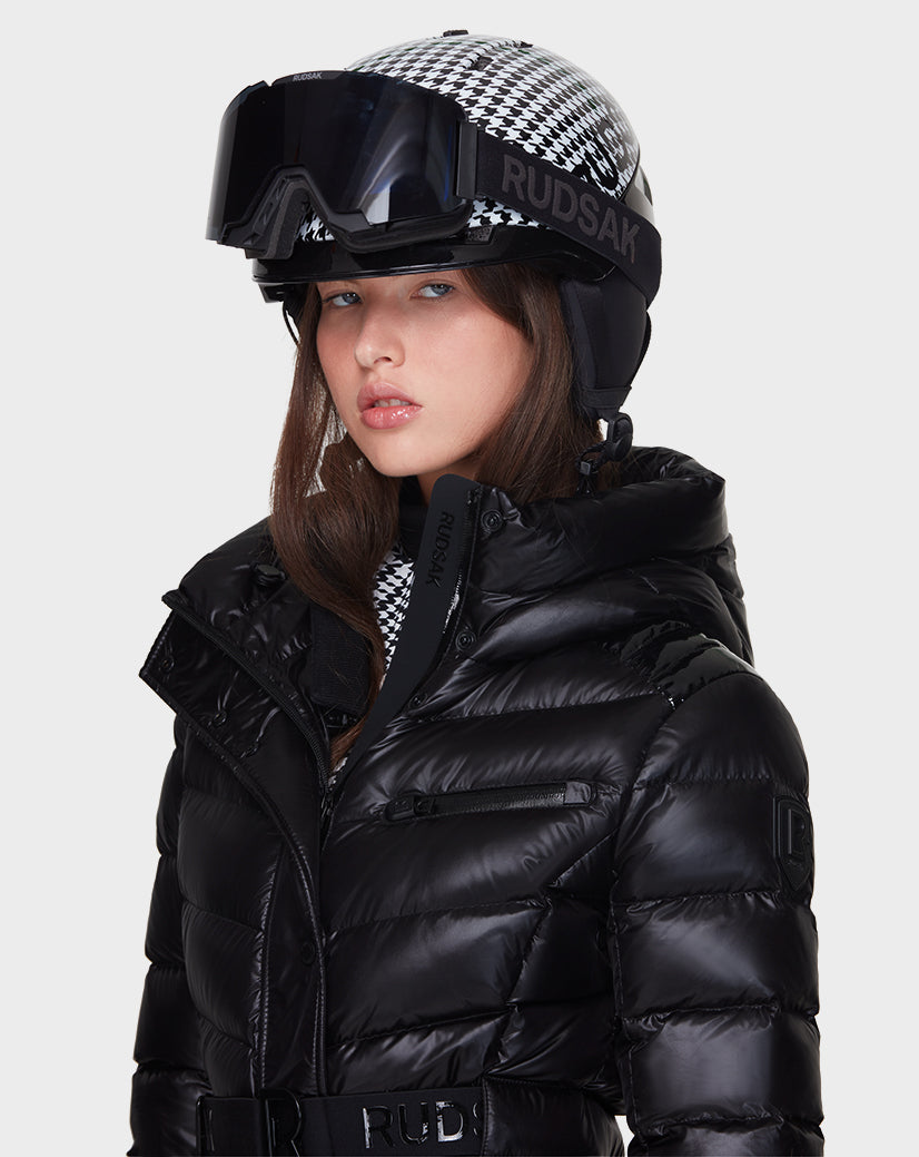 A woman wearing the Rudsak MASORA - 8121535 BLACK shiny down puffer jacket and a black-and-white houndstooth ski helmet with attached goggles, both branded Rudsak.