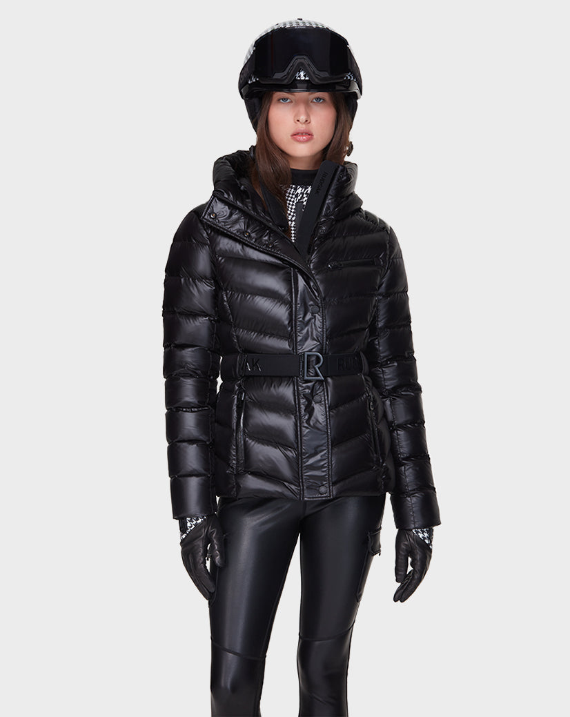 A woman wearing the Rudsak MASORA - 8121535 BLACK shiny down ski jacket with matching black helmet, goggles, gloves, and pants stands facing forward against a plain white background.
