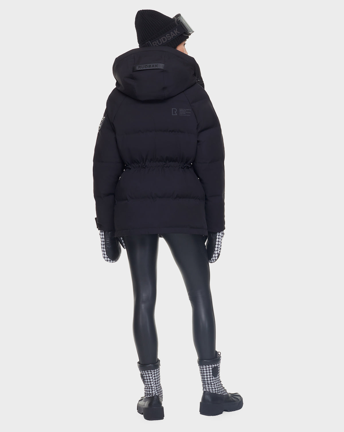 A person wearing the Rudsak MANDORA - 8121542 BLACK women’s down parka with a hood, plus a black beanie, black leggings, houndstooth gloves, and matching boots stands facing away against a plain light background.