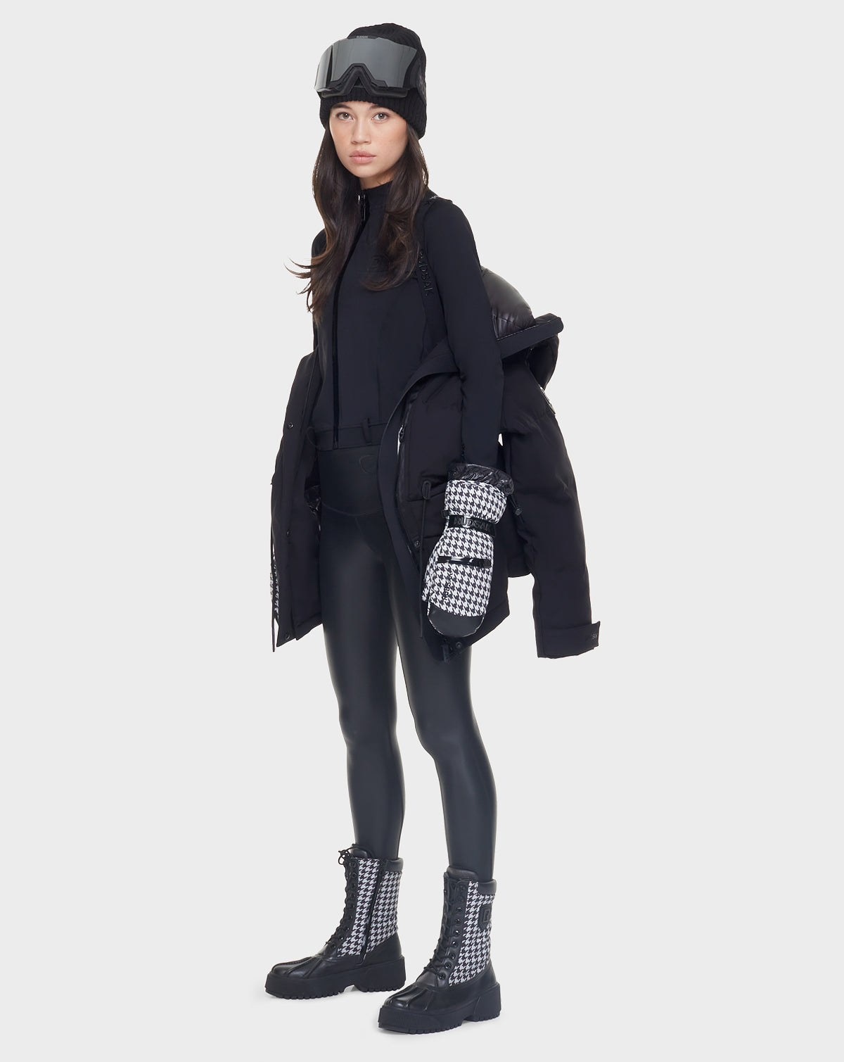 A woman faces forward against a plain background, wearing black ski goggles, a black knit hat, checkered mittens and boots, shiny black leggings, a black turtleneck, and the Rudsak MANDORA - 8121542 BLACK women’s down parka draped over her shoulders.