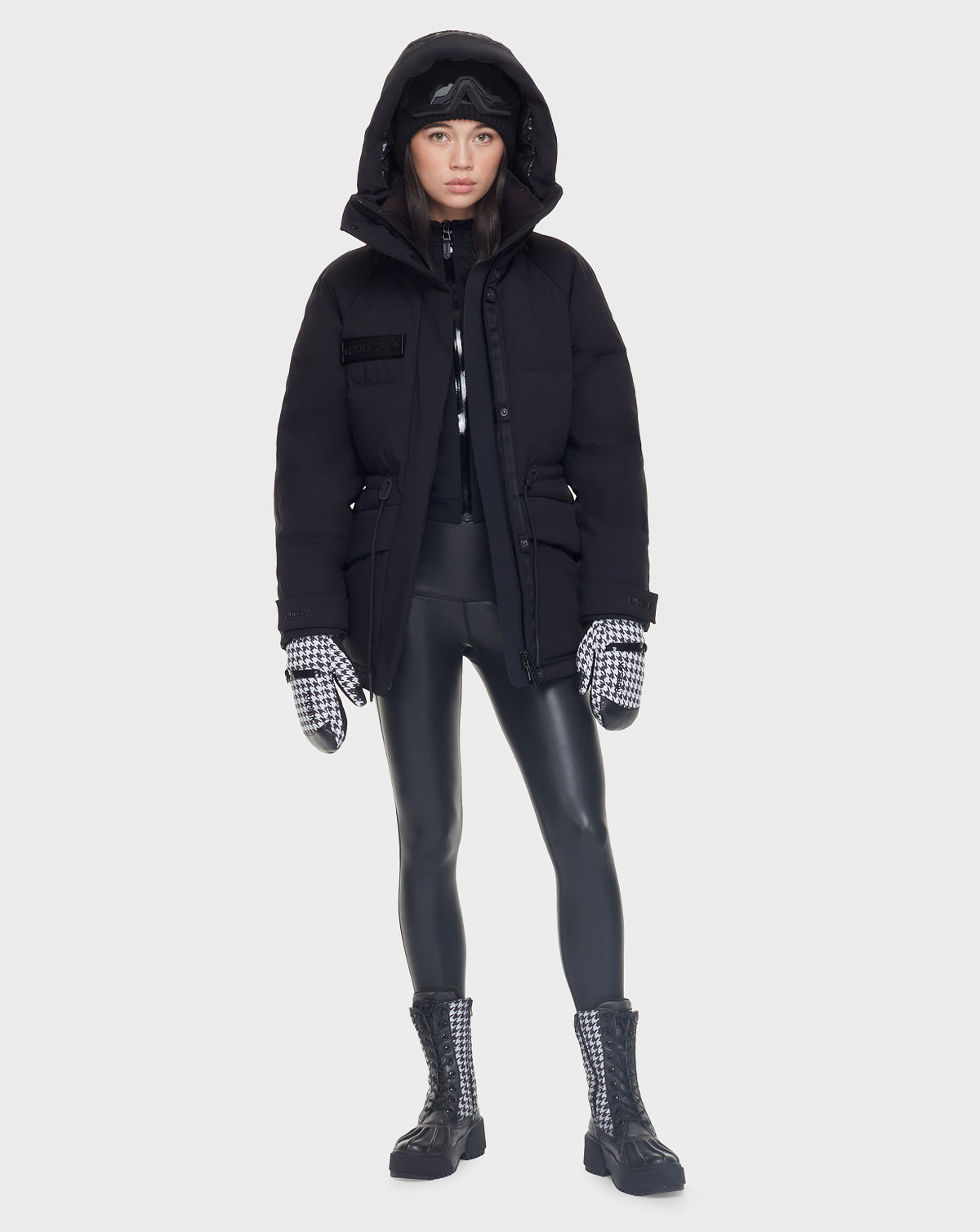 A person wearing the Rudsak MANDORA - 8121542 BLACK parka, black leggings, plaid gloves and boots, a black knit hat, and ski goggles stands facing forward against a plain white background.