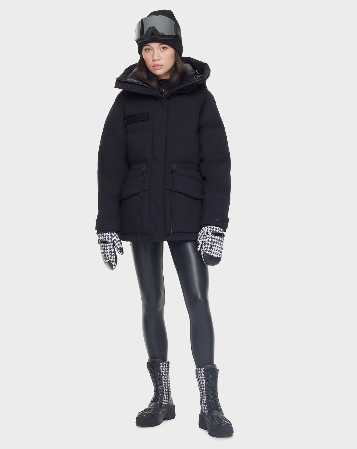 A woman stands facing forward against a plain light background, wearing the Rudsak MANDORA - 8121542 BLACK womens down parka, shiny black leggings, black boots, houndstooth gloves, and ski goggles.